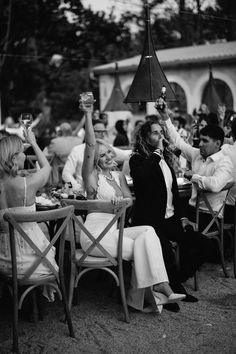 black and white photograph of people sitting at tables with drinks in their hands, one woman raising her hand up to the sky