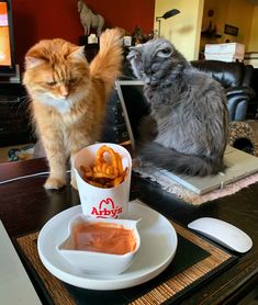 two cats sitting at a table with food on it and one cat standing next to them