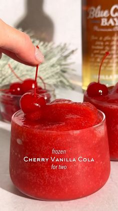 a hand is holding a cherry vanilla cola in front of two small jars with cherries on them