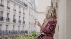 a woman standing on a balcony holding a glass of wine in her hand and looking out the window