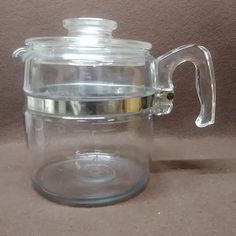 a clear glass teapot with a lid on a brown tableclothed surface next to a wall