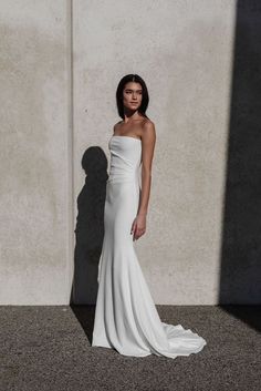 a woman standing in front of a wall wearing a white dress with one shoulder open