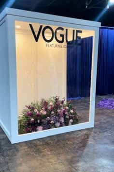 a display case with flowers in it on the floor and blue curtains behind it that says vogui