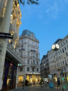 people walking on the street in front of some tall buildings with clocks mounted to it's sides