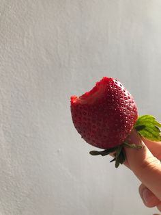 a hand holding a strawberry with green leaves on it's tip and white wall in the background