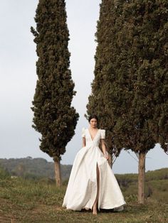 a woman in a white wedding dress standing next to two tall trees on a hill