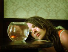 a woman laying her head on a table next to a fish bowl