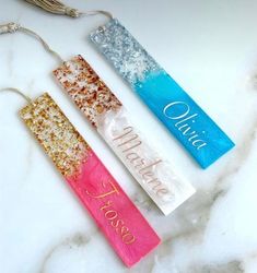 three personalized bookmarks with tassels hanging from them on a marble surface