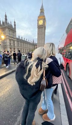 two women are walking down the street in front of big ben, with a red double decker bus behind them