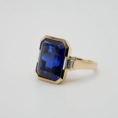 This exquisite 12 Ct Lab Grown Emerald Cut Sapphire and Trapezoid Diamond Ring is masterfully set in 18K yellow gold. This piece captures the essence of modern elegance with its sharp, clean lines and a striking bezel setting. Flanked by trapezoid diamonds, the sapphire centerpiece exudes a royal blue hue. Perfect for those who cherish contemporary design with a hint of classic allure. Emerald Cut Rings, Three Stone Ring, Three Stone Rings, Modern Elegance, Three Stone, Blue Hues, Stone Ring, Bezel Setting, Emerald Cut