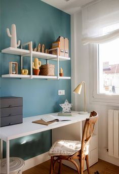 a white desk sitting under a window next to a wooden chair and shelf filled with books