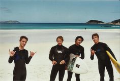 three men in wetsuits standing on the beach with their surfboards and making peace signs