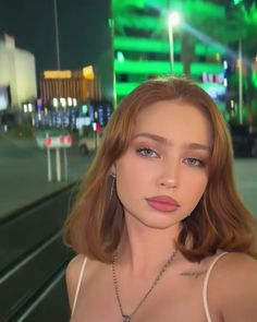 a woman with red hair wearing a white top and silver necklace standing in front of a city street at night