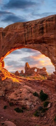 an arch shaped rock formation with the sun setting in the background and clouds above it