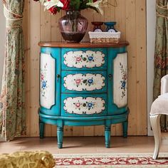 a blue cabinet with flowers on it in front of a wooden paneled wall and curtains