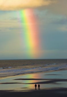 two people standing on the beach under a rainbow colored cloud over the ocean and water