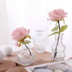 three glass vases with flowers in them on a wooden tray next to rocks and eggs