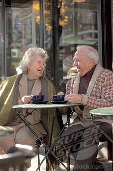 two elderly people sitting at an outdoor cafe talking to each other and drinking tea or coffee