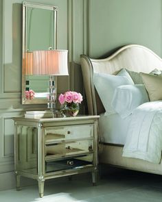 a white bed sitting next to a mirror and lamp on top of a wooden table