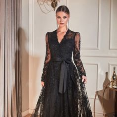 a woman wearing a black dress with long sleeves and a bow at the waist, standing in front of a chandelier