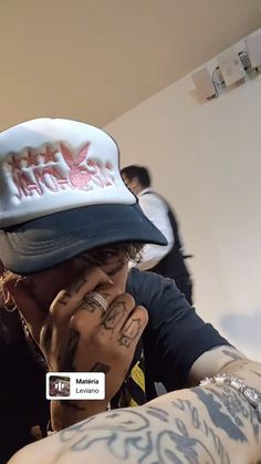 a man with tattoos covering his face and holding his hands up to his mouth while wearing a hat