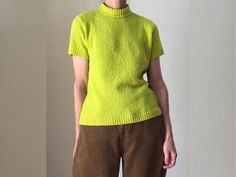 a woman standing in front of a white wall wearing a yellow sweater and brown pants