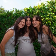 three women standing next to each other in front of some bushes and trees with one woman smiling at the camera