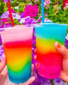 two people holding up colorful drinks in front of purple and pink flowers with straws