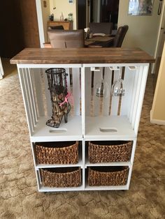 a white cabinet with two wine glasses on top and some baskets underneath the doors, in a living room