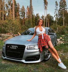 a beautiful woman leaning on the hood of a silver car in front of some trees