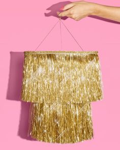 a woman's hand holding a gold fringe lamp shade on a pink background,