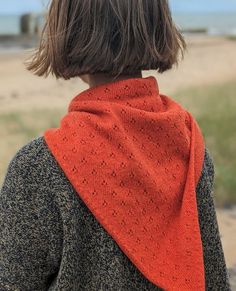 a woman wearing an orange knitted shawl on top of a sandy beach next to the ocean