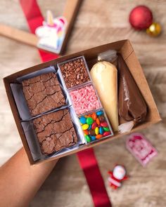 a person holding a box filled with different types of candy and brownies on top of a wooden table