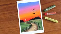 some crayons are sitting on a table next to a card with an image of a sunset