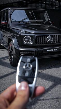 a hand holding a remote control in front of a black mercedes g - class car