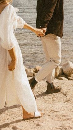 a man and woman holding hands while walking on the sand near water's edge