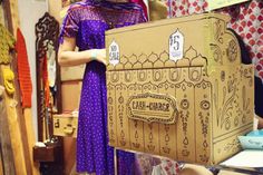 a woman standing next to a cardboard box in a room filled with clothes and other items