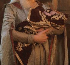 a man with long white hair wearing a brown and gold outfit holding a baby in his arms