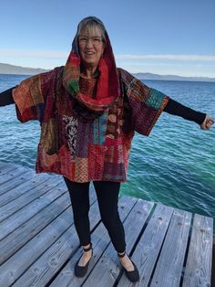 an older woman standing on a dock with her arms out in the air and wearing a colorful scarf over her head