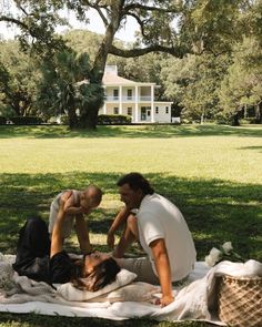 two people sitting on a blanket in front of a white house with trees and grass