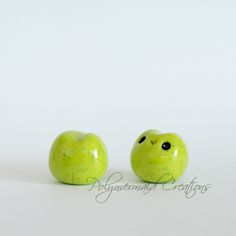 two green apples sitting side by side on a white surface, one with black eyes and the other has yellow speckles