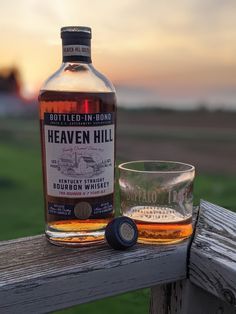 a bottle of heaven hill whiskey next to a shot glass on a wooden rail with the sun setting in the background