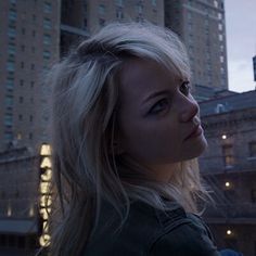 a woman with blonde hair standing in front of tall buildings at night, looking off into the distance