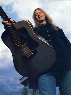 a woman holding an acoustic guitar in front of a blue sky with clouds behind her