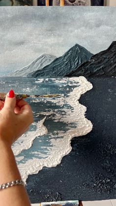 a person is holding a paintbrush and painting an ocean scene with mountains in the background