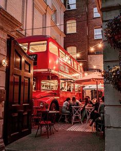 a red double decker bus parked in front of a building with people sitting at tables