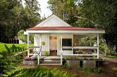 a small white house with a red roof in the middle of some trees and bushes