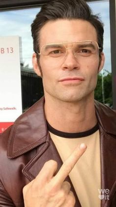 a man with glasses and a leather jacket pointing to the side while standing in front of a window