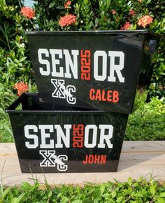 two black containers with red and white letters are sitting on the ground in front of some bushes