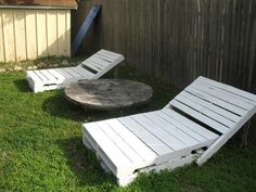 two white lounge chairs sitting in the grass next to a wooden table and fenced in area